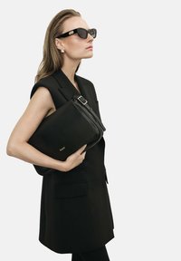 Borsa in pelle nera tenuta sotto braccio, caratterizzata da una fibbia argentata e un logo in rilievo. Il modello indossa un gilet nero e occhiali da sole con montatura spessa.