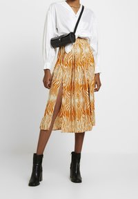 Jupe midi à motifs orange et crème avec des fentes latérales, associée à une chemise blanche boutonn ée et des bottines noires. Sac bandoulière noir.