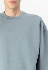 Lichtblauwe sweatshirt met een geribde ronde hals, verlaagde schouders en een reliëf "Hollister" logo aan de voorkant. Zachte stof structuur.