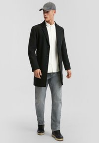 Schwarzer langer Mantel, weißes Hemd, graue Jeans und schwarze Sneakers; das Model trägt eine graue Mütze. Glattes Material mit Knöpfen und taillierter Passform.