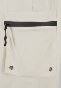 Taschina in tessuto beige chiaro con zip nera e linguetta, dotata di due occhielli metallici agli angoli inferiori per rinforzo.