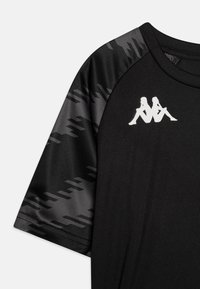 Černé atletické tričko s texturovaným černým vzorovaným rukávem a bílým logem na hrudi. Materiál působí hladce a lehce.