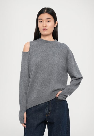 Maglione grigio con design a spalla scoperta, polsini a coste e vestibilità rilassata, abbinato a jeans blu scuro, larghi. Texture liscia.