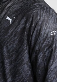 Chaqueta negra con un patrón texturizado en tonos de gris. Cuenta con un logo blanco de Puma y sutiles puntos reflectantes en el hombro.