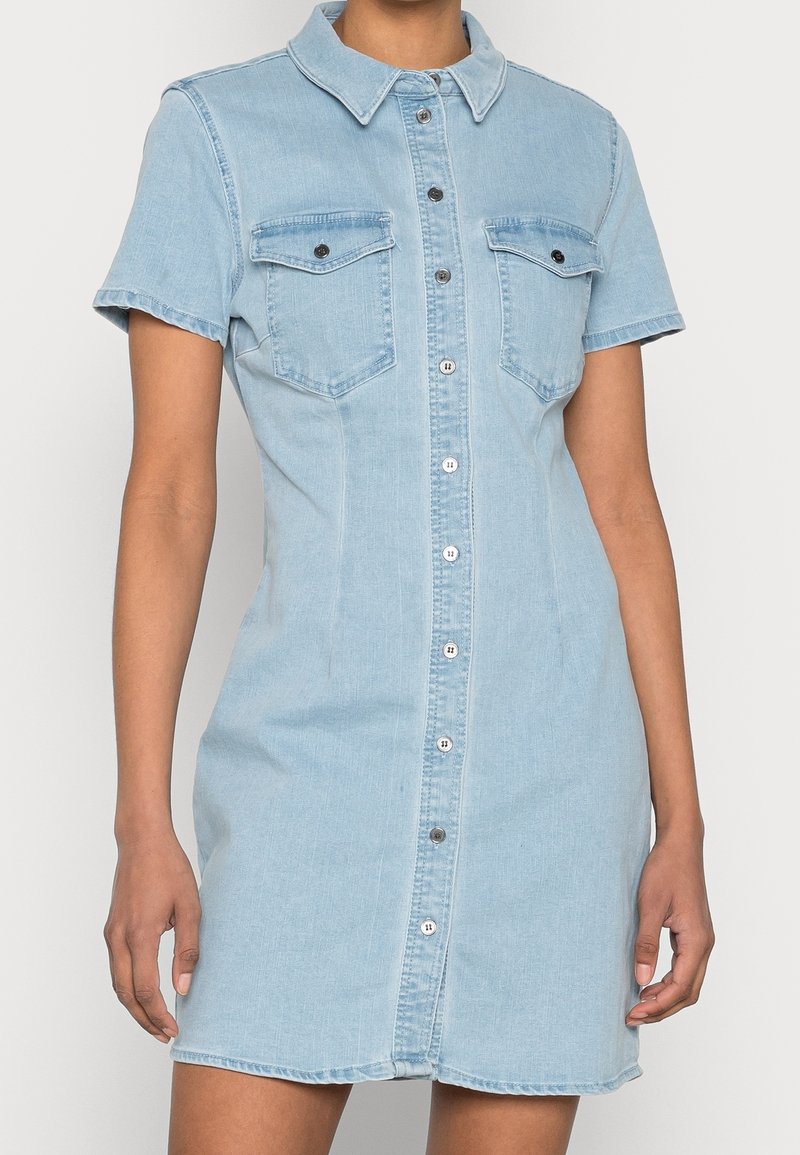 Robe chemise en denim bleu clair avec manches courtes, fermeture à boutons devant, deux poches poitrine et une taille cintrée. Texture lisse et col.