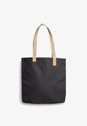Sac fourre-tout noir avec des anses d'épaule beiges et une petite étiquette de marque orange sur le côté supérieur droit, sur fond blanc.