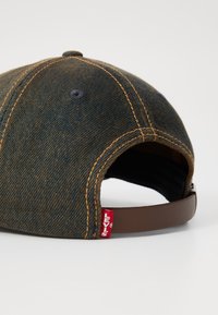Denim kapa z temno modrim in rjavim barvnim vzorcem, ki vključuje oranžno šivanje, nastavljiv rjav trak in rdečo oznako Levi's.