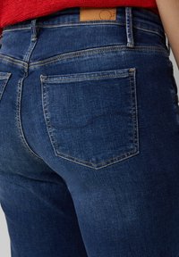 Donkerblauwe denim jeans met een close-up van de achterkant, met contrasterende gele stiksels en een bruine leren patch op de tailleband.