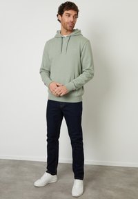 Lichtgroene hoodie van zacht materiaal met een buidelzak en verstelbare koordjes, gecombineerd met donkerblauwe jeans en witte sneakers.