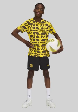 Maglietta da calcio con motivo geometrico giallo e marrone, pantaloni corti neri con logo, pallone da calcio bianco con dettagli verdi, calze bianche e scarpe da calcio.