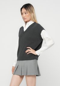 Maglione grigio a collo a V senza maniche, indossato sopra una camicia bianca con colletto, abbinato a una mini gonna plissettata grigia.