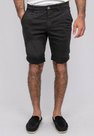Homme portant un short noir retroussé, une chemise blanche, des chaussures noires sans lacets, les mains relâchées le long du corps devant un fond clair uni.