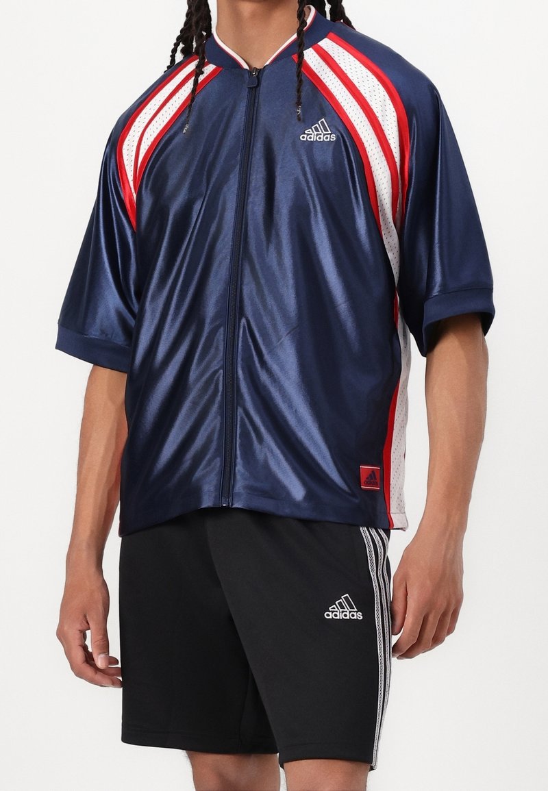 Námořnická zipová sportovní košile s červenými a bílými síťovanými akcenty, krátkými rukávy a lesklou texturou. Na hrudi a na lemu se nachází logo Adidas.