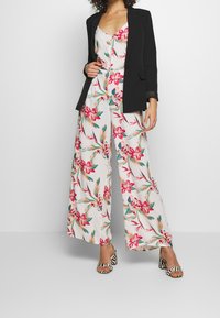 Combinaison florale avec des motifs roses et verts sur fond blanc, associée à un blazer noir et des sandales à talons rayées.