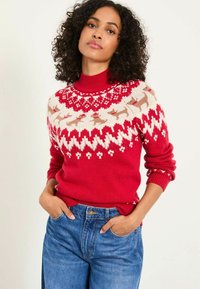 Frau mit lockigem Haar, die einen roten Rollkragenpullover mit einem weißen und beigen Rentiermotiv trägt, mit den Händen in den Taschen ihrer blauen Jeans.