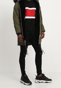 Parka verde oliva con foderatura in pelliccia sintetica nera, felpa nera con una grafica rossa e bianca, jeans neri strappati, sneakers nere.
