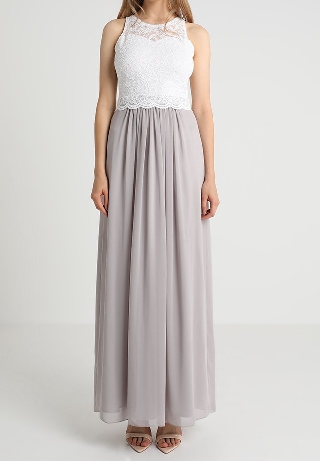 Femme portant un haut en dentelle blanche sans manches avec un col montant et une longue jupe fluide gris clair ainsi que des sandales beiges.