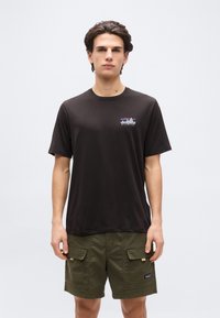 Νεαρός άνδρας στέκεται με το πρόσωπο προς τα μπροστά, φοράει μαύρο μπλουζάκι Patagonia και χακί cargo σορτς, μπροστά από ένα απλό φόντο.