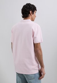 Mężczyzna z krótkimi kręconymi włosami, ubrany w jasnoróżową koszulkę polo z krótkim rękawem i jasnoniebieskie dżinsy, zwrócony plecami na jednolitym tle.