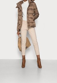 Giacca puffa marrone con cappuccio, pantaloni beige attillati, stivaletti marroni e una borsa leggera color beige, il tutto in una palette di colori neutri.