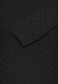Sort strikket sweater med diamantmønster og ribbede ærmer, lagt over brystområdet.