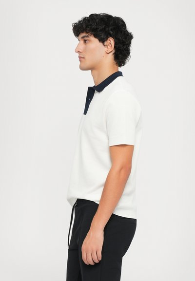 Kortermet polo skjorte i hvit bomull med marineblå krage og knappeåpning. Modellen har på seg svarte bukser og viser en sideprofil.