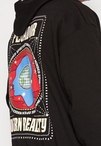 Schwarzer Kapuzenpullover mit einem bunten Grafikdruck auf der Rückseite, der einen Globus mit roten und blauen Akzenten sowie stilisierten Textelementen darstellt.