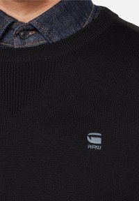 Schwarzer Strickpullover mit Rundhalsausschnitt, gerippter Struktur und einem kleinen blauen Logo auf der Brust. Darunter befindet sich ein Jeanshemd mit knöpfbarem Kragen.