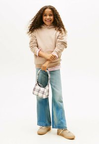 Chica sonriente con cabello largo y rizado, vistiendo un sudadera beige, una camiseta a rayas, jeans de pierna ancha y zapatillas beige, sosteniendo un bolso de cuadros, todo sobre un fondo blanco.