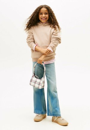 Chica sonriente con cabello largo y rizado, vistiendo un sudadera beige, una camiseta a rayas, jeans de pierna ancha y zapatillas beige, sosteniendo un bolso de cuadros, todo sobre un fondo blanco.