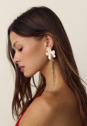 Woman with long brown hair wearing a red top and large floral earring with gold chains and small white pearls, facing left in neutral background.