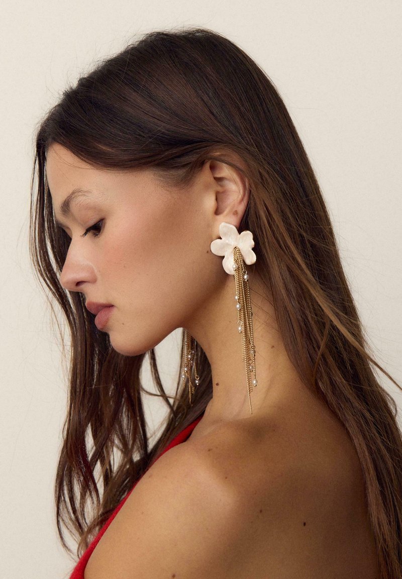 Femme aux longs cheveux bruns portant un haut rouge et une grande boucle d'oreille florale avec des chaînes en or et de petites perles blanches, tournant le visage vers la gauche sur un fond neutre.