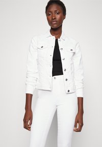 Jaqueta de ganga branca com dois bolsos no peito e botões pretos, usada sobre uma blusa preta, combinada com calças brancas, contra um fundo simples.