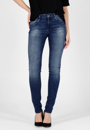 Slim fit blauw vervaagde jeans gedragen met zwarte hoge hakken en een zwarte top, onderlichaam en benen zichtbaar, staand tegen een effen achtergrond.