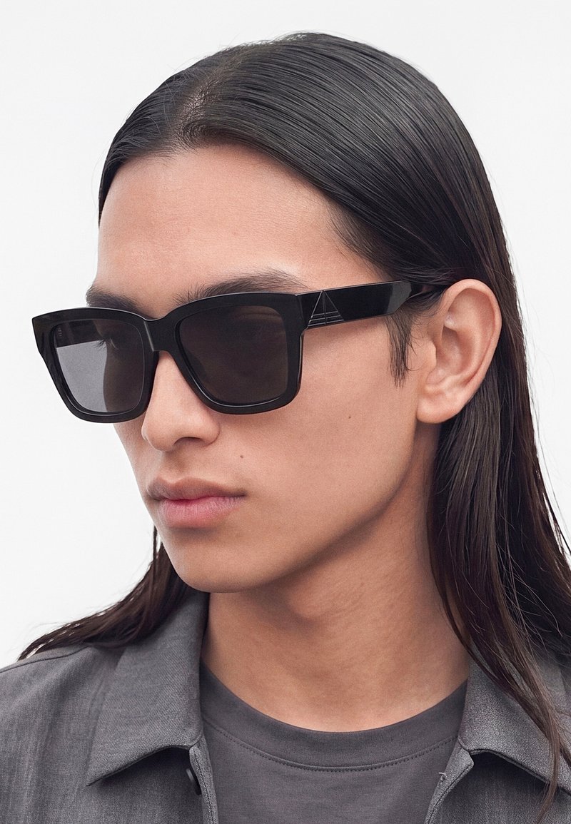 Jeune personne aux longs cheveux foncés portant de grandes lunettes de soleil carrées noires et une chemise et une veste grises, regardant sur le côté.