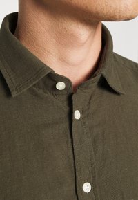 Camisa verde oliva de botones con cuello doblado, botones blancos y un acabado de tela texturizada, mostrando detalles de costura cerca de los botones.