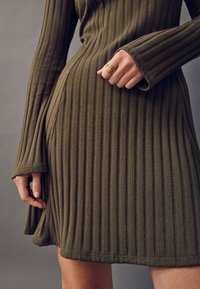 Das olivgrüne, gerippte Kleid verfügt über lange Ärmel und einen ausgestellten Saum mit seitlichem Schlitz und ordentlicher Nahtverarbeitung. Ein goldener Statement-Ring wird getragen.