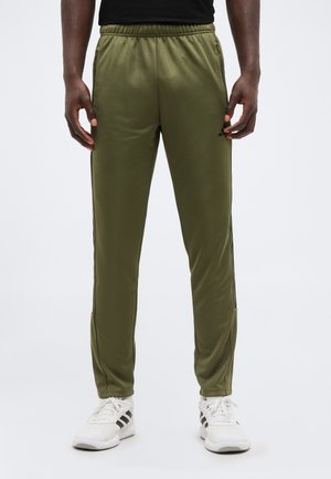 Mężczyzna model ubrany w oliwkowozielone sportowe spodnie z paskami po bokach i białe trampki, stojący na tle jednolitego jasnego tła.