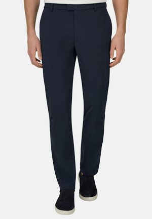 Mann trägt dunkelblaue Slim-Fit-Hose und schwarze Slipper mit weißen Sohlen, steht vor weißem Hintergrund.