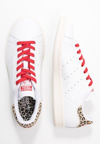 Weiße Sneakers mit roten Schnürsenkeln, perforiertem Design und einem beigen Leopardenmusterakzent an der Ferse. Weiches Leder mit einer strukturierten Sohle.