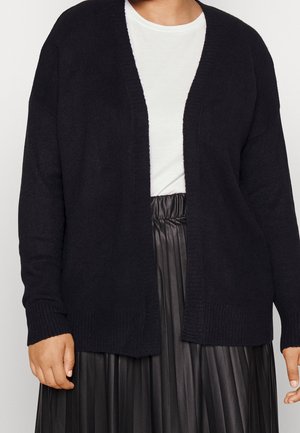 Person iført en sort cardigan med åben front, hvid T-shirt med rund hals og sort plisseret nederdel med høj talje, vist fra skuldre til talje.