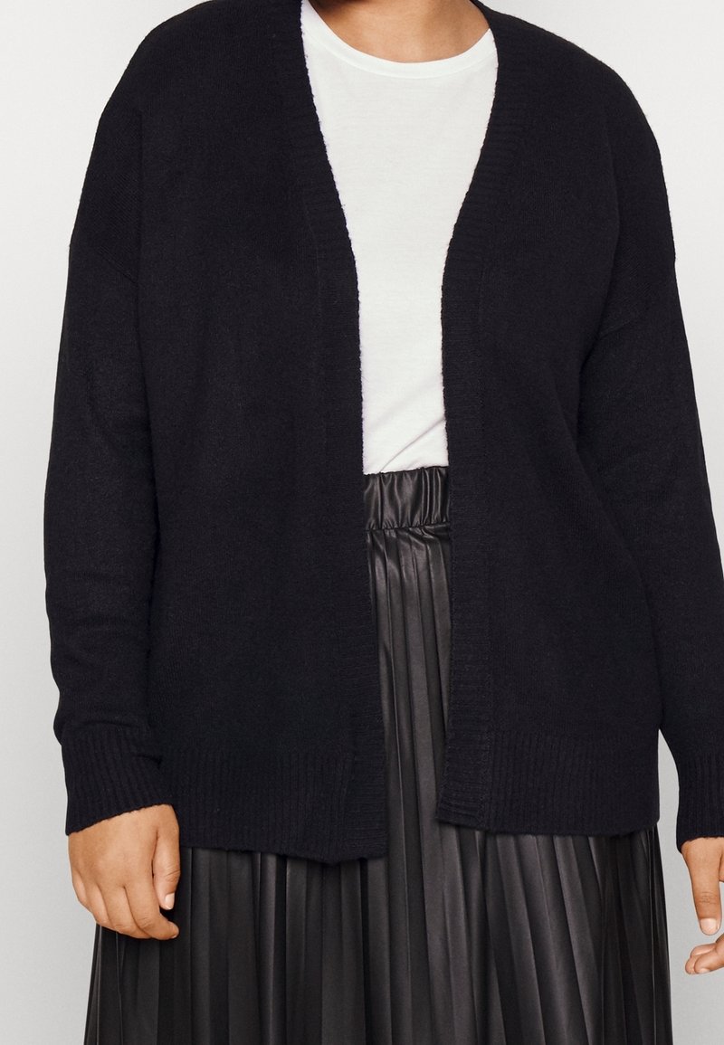 Personne portant un cardigan noir ouvert devant, un t-shirt blanc à col rond et une jupe plissée noire taille haute, vue des épaules à la taille.