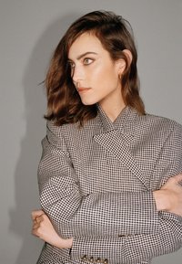 Blazer à carreaux en marron et crème, avec un devant double boutonnage, des épaules structurées et un tissu texturé agrémenté de boutons dorés.