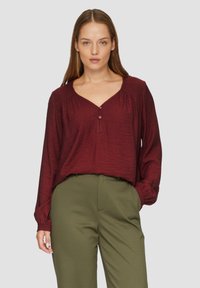 Blouse bordeaux à manches longues avec un col en V, dotée d'un tissu texturé et de deux boutons au col. Associée à un pantalon vert olive.