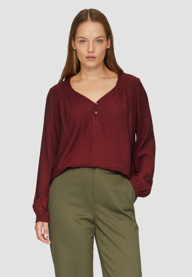 Blouse bordeaux à manches longues avec un col en V, dotée d'un tissu texturé et de deux boutons au col. Associée à un pantalon vert olive.