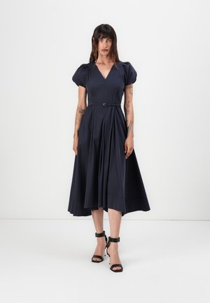 Navy blue dress with a V-neck, puff sleeves, and a belted waist. Pleated skirt extends below the knee. Worn with open-toe black heels.