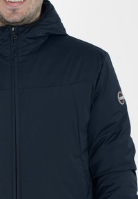 Veste matelassée bleu marine avec capuche, dotée d'une fermeture éclair et d'un écusson sur la manche gauche. Texture lisse et design léger.
