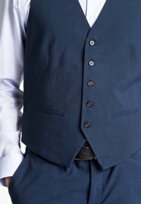 Navyblaues Weste aus glattem Stoff, mit sechs Knopfverschlüssen, zwei Seitentaschen und einem spitzen Saum. Kombiniert mit einem hellblauen Hemd.