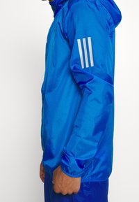 Mężczyzna w jasnoniebieskiej kurtce z kapturem z trzema białymi paskami na górnej części rękawa, stojący na prostym tle.