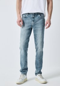 Vaqueros de mezclilla de color azul claro con corte recto, que presentan cinco bolsillos y un leve desgaste. Llevados con zapatillas blancas y una camiseta blanca lisa.